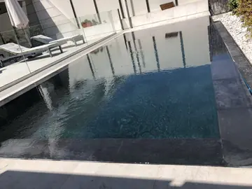 Piscina con borde gris oscuro. Piscina con revestimiento gris oscuro, borde a ras del agua y playa en tono crema junto a la barandilla de vidrio.