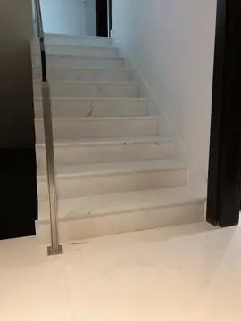 Tramo de escalera con poste de acero. Peldaños blancos con veta gris, zanquín a juego y barandilla con poste de acero sobre suelo pulido.