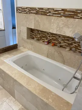 Bañera con frente y hornacina revestidos. Bañera encastrada con frente y repisa en piezas beige y hornacina con fondo de mosaico en marrón, crema y gris.