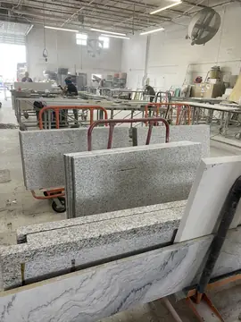Piezas cortadas en caballetes del taller. Piezas pulidas moteadas en gris y blanco y un tablero de veta gris en caballetes, con el taller trabajando al fondo.