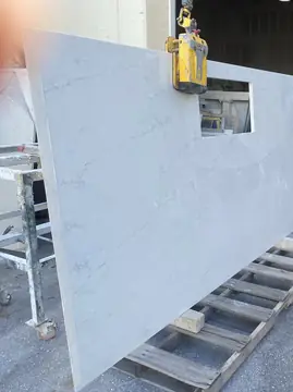Encimera izada con pinza de elevación. Encimera blanca con veta gris y hueco rectangular, levantada con una pinza de elevación sobre un palé.