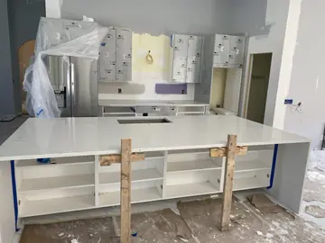 Isla con puntales frente a la cocina. Encimera blanca de la isla sujeta con puntales de madera, con los muebles bajos abiertos y la cocina al fondo.