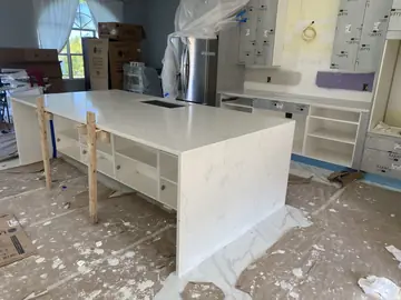 Isla con faldón y cocina en montaje. Isla con encimera blanca de veta suave y faldón hasta el suelo, con la bancada y los muebles altos en montaje al fondo.