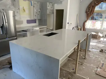 Isla colocada en cocina en obra. Isla con encimera blanca de veta gris y faldón hasta el suelo, con la bancada y los muebles altos al fondo.