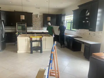 Operarios llevando una encimera a su sitio. Dos operarios de espaldas llevan una pieza de encimera hacia la bancada de una cocina de muebles oscuros.