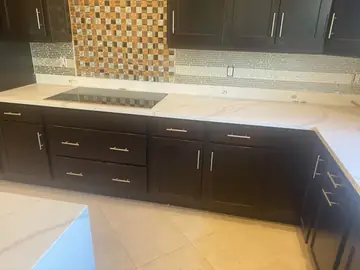 Bancada con placa y frontal de mosaico. Encimera blanca con veta gris sobre muebles oscuros, con placa de cocción y frontal de mosaico.