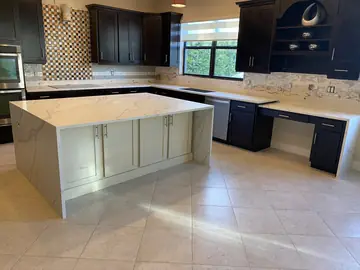 Cocina con isla y bancada perimetral. Vista de la cocina con isla de faldones laterales y bancada perimetral en encimera blanca de veta gris.