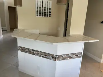 Barra con encimera y franja de mosaico. Barra de salón con encimera blanca de veta gris y frente revestido con pieza en relieve y franja de mosaico.