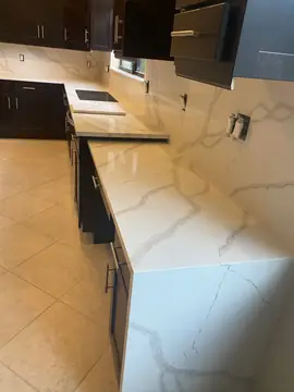 Bancada con faldón y frontal a juego. Encimera con faldón hasta el suelo y frontal de pared en piezas blancas con veta gris sobre muebles oscuros.