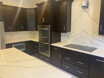 Bancada con placa y frontal hasta arriba. Encimera y frontal blancos con veta gris junto a la placa de cocción, con hornos en columna y muebles oscuros.