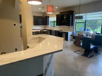 Barra y cocina vistas desde el salón. Barra con encimera blanca de veta gris en primer plano y la cocina con isla y frontal veteado al fondo.