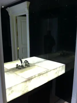 Lavabo retroiluminado frente al espejo. Encimera con lavabo integrado en placa translúcida iluminada desde dentro, ante un espejo y paredes oscuras con veta blanca.