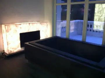 Chimenea retroiluminada junto a la bañera. Frente de chimenea en placas translúcidas iluminadas desde dentro junto a una bañera revestida en tono oscuro ante el ventanal.