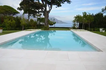 Piscina con pavimento claro frente al mar. Piscina rodeada de pavimento en piezas claras de gran formato, con jardín y el mar al fondo.