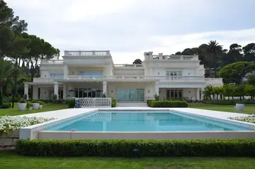 Piscina y jardín ante la casa. Piscina con borde claro en el jardín, delante de una casa de estilo clásico con balaustradas.