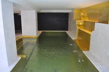 Piscina cubierta con paredes claras. Piscina interior con paredes y bordes en tono claro y un paño de mosaico dorado en el lateral.