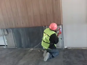 Remate del zócalo oscuro con pistola. Un operario trabaja con pistola de cartucho en el hueco junto al zócalo oscuro, bajo paneles en tono madera.