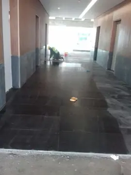 Pavimento negro en pasillo con zócalo oscuro. Pavimento negro en piezas grandes a lo largo del pasillo, con zonas mojadas y un operario trabajando al fondo.