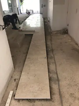 Replanteo de la primera hilera del pavimento. Primera franja de piezas en tono crema asentada sobre la solera, con operarios avanzando la colocación al fondo.