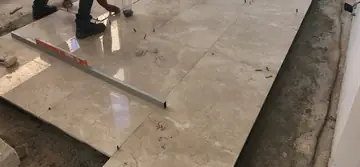 Colocación de piezas grandes de pavimento. Operarios nivelan una pieza pulida en tono crema junto a la franja ya asentada, con crucetas y regla de aluminio.