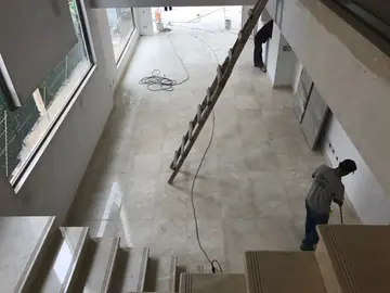 Doble altura con pavimento y escalera. Salón visto desde la escalera revestida, con pavimento pulido en tono crema y operarios terminando la limpieza.