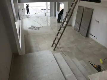 Salón en doble altura con pavimento colocado. Pavimento pulido en tono crema y arranque de escalera revestido, con escalera de mano y cables de la obra.