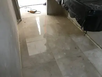 Pavimento pulido en un pasillo en obra. Piezas en tono crema con brillo y veta hasta la puerta, junto a un volumen cubierto con plástico negro y paredes aún sin terminar.