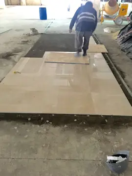 Colocación de pavimento pulido en tono crema. Piezas grandes de pavimento pulido en tono crema recién asentadas sobre el mortero, con un operario caminando sobre un tablero al fondo.