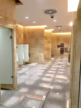 Aseo público revestido en tono crema. Zona de urinarios con paredes y pilastras revestidas en tono crema, iluminación indirecta y suelo gris moteado con banda central.