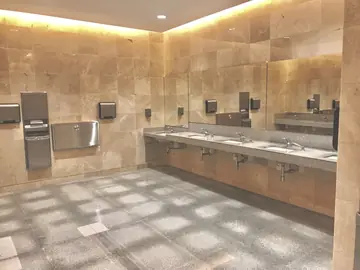 Lavabos corridos en aseo público. Aseo con paredes revestidas en piezas tono crema con vetas, encimera corrida gris moteada con lavabos y pavimento gris.