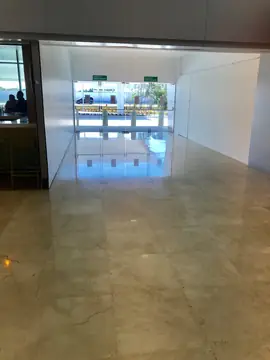 Paso hacia salida con suelo en tono crema. Pasillo con pavimento pulido en tono crema que refleja la luz de las puertas de vidrio de salida al exterior.