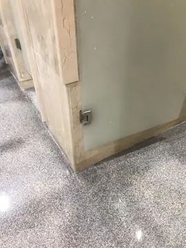 Encuentro de jamba y pavimento. Detalle de jamba revestida en tono crema con herraje de puerta de vidrio sobre pavimento pulido gris moteado.