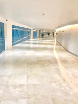 Corredor acristalado con suelo pulido. Corredor con pavimento pulido en tono crema, frente de vidrio azulado a la izquierda y luz indirecta en la pared derecha.