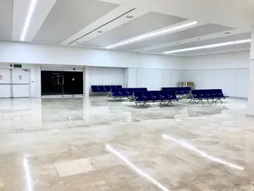 Sala de espera con suelo en tono crema. Sala de espera con bancadas azules sobre pavimento pulido en tono crema con vetas y techo de luminarias lineales.