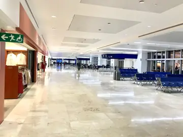 Zona de embarque con pavimento pulido. Gran zona de espera con pavimento pulido en tono crema, tiendas a la izquierda y bancadas azules a la derecha.
