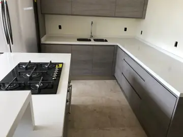 Cocina en U con isla y placa de gas. Encimeras blancas en la isla y en el tramo del fregadero, con muebles grises de veta tipo madera.