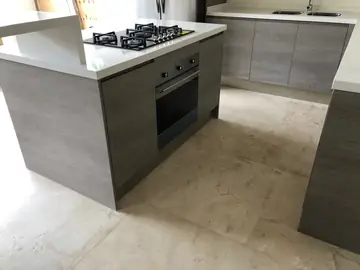 Isla con horno y placa frente a la cocina. Isla con encimera blanca, placa de gas y horno integrado, frente al tramo del fregadero y sobre pavimento en tono crema.