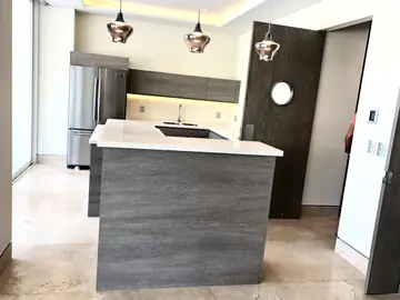 Cocina con península y lámparas colgantes. Península con encimera blanca y frente gris de veta tipo madera, con la cocina al fondo y pavimento pulido en tono crema.