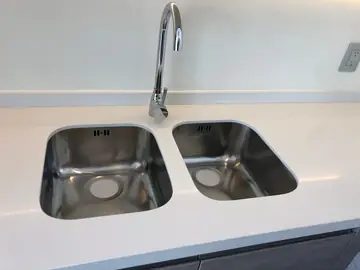 Fregadero doble bajo encimera. Fregadero doble de acero bajo encimera blanca, con grifo de caño alto y copete a lo largo de la pared.