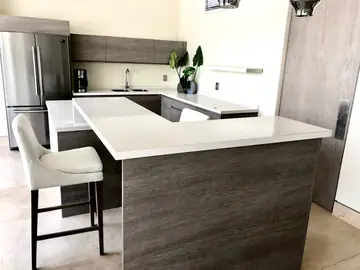 Cocina terminada con península y taburete. Península con encimera blanca y frente gris de veta tipo madera, abierta a la cocina en U con frigorífico de acero.
