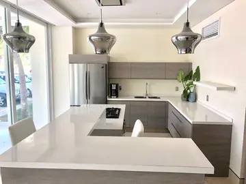 Cocina con isla bajo lámparas colgantes. Isla con encimera blanca, placa de gas y zona de barra, frente a la cocina en U con muebles grises.