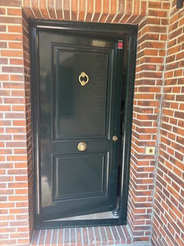 Puerta de entrada en verde oscuro. Lacada a brillo en verde muy oscuro, con el cerco del mismo color, aldaba de argolla y pomo de latón, enmarcada en el ladrillo.