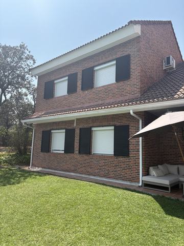 Fachada de ladrillo visto. Esquina de la vivienda con contraventanas negras y jardín a pleno sol.