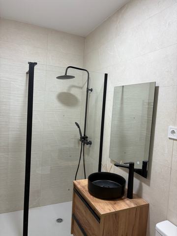 Baño con ducha a ras de suelo y lavabo negro. Ducha a ras de suelo con plato blanco extraplano, mampara fija de cristal con perfil negro y columna de ducha negra. Mueble suspendido de dos cajones en efecto roble con lavabo redondo negro sobre encimera y grifo alto negro. Alicatado en porcelánico beige.