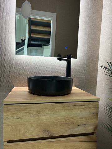 Lavabo negro y espejo LED. Mueble de dos cajones en acabado efecto roble, lavabo redondo negro apoyado sobre la encimera, grifo alto negro y espejo con luz LED.