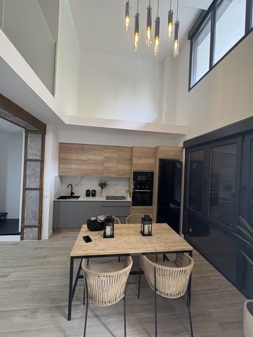 Cocina abierta al comedor de doble altura. Cocina en línea al fondo, bajo falso techo rebajado con focos empotrados: altos y columna en acabado madera con veta, bajos en gris mate, encimera y frente con veta tipo mármol, fregadero y grifo negros, placa de inducción y horno con microondas en negro. Por delante, el comedor bajo la doble altura, con seis lámparas de tubo colgadas del techo alto y la carpintería negra acristalada a la derecha.