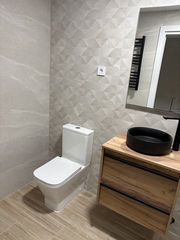 Baño en gris y roble. Porcelánico gris de gran formato: relieve geométrico en la pared del fondo y liso con veta en el lateral. Suelo de porcelánico imitación madera, mueble de dos cajones en acabado efecto roble con tiradores negros y lavabo redondo negro sobre encimera.