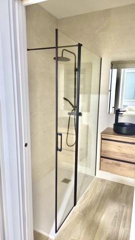 Ducha con mampara y grifería negras. Ducha con mampara de vidrio y perfilería negra, puerta abatible con tirador, columna con rociador de lluvia y teléfono, y plato blanco con sumidero cuadrado. Alicatado porcelánico beige de gran formato y suelo imitación tarima. A la derecha, mueble suspendido de dos cajones en acabado madera, con lavabo redondo negro sobre encimera y grifo negro.