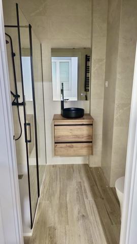 Baño alargado con ducha a ras de suelo. Ducha a ras de suelo con mampara de vidrio de perfil negro y columna de ducha negra. Mueble suspendido de dos cajones en roble con encimera de madera, lavabo redondo negro y grifería mural negra. Paredes en porcelánico beige de gran formato y suelo porcelánico imitación madera.